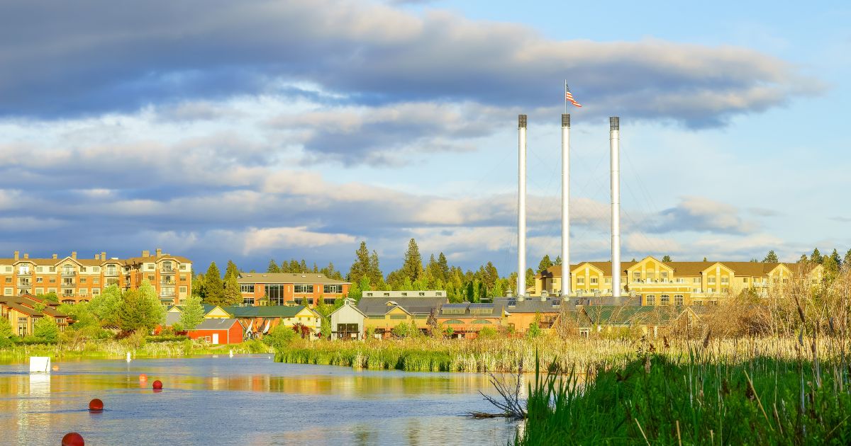 Waterfront town with buildings, tall smokestacks with American flag, and green vegetation under a cloudy sky.